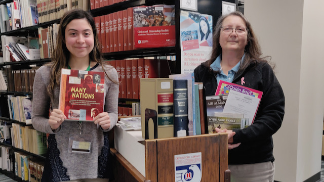 Two women in front of book shelves holding up government publications.